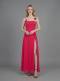 Woman in a bright pink ruched maxi dress with spaghetti straps and butterfly details, standing against a grey backdrop.