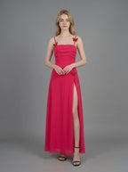 Woman in a bright pink ruched maxi dress with spaghetti straps and butterfly details, standing against a grey backdrop.