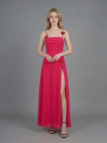 Woman in a bright pink ruched maxi dress with spaghetti straps and butterfly details, standing against a grey backdrop.