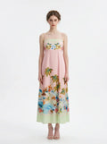 Woman wearing a pink midi dress with spaghetti straps and a tropical print of palm trees and flowers.