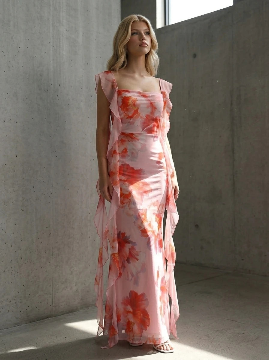 Woman posing in a pink floral print chiffon maxi dress with ruffle trim against a concrete wall