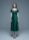 Woman wearing a green satin midi dress with puff sleeves and a square neckline, standing against a gray background.