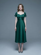 Woman wearing a green satin midi dress with puff sleeves and a square neckline, standing against a gray background.