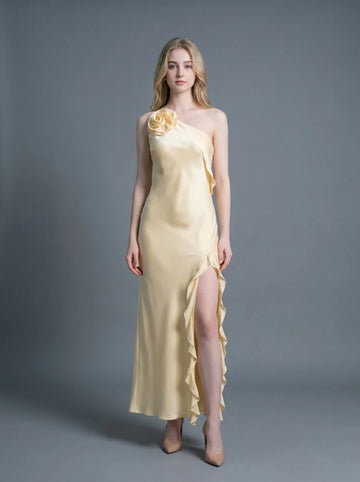 Woman wearing a pale yellow satin one-shoulder gown with a ruffled high slit and a floral applique on the shoulder.
