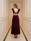 Back view of a woman in a burgundy velvet burnout A-line maxi dress with an open back and lace-up detail, standing in a hallway.