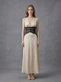 Woman wearing a cream satin maxi dress with black lace detailing at the waist and a halter neckline, standing against a grey backdrop.