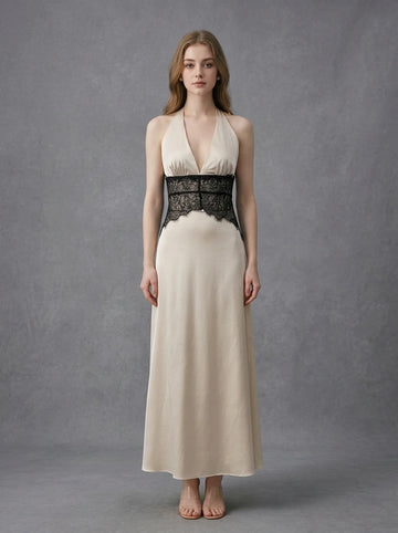 Woman wearing a cream satin maxi dress with black lace detailing at the waist and a halter neckline, standing against a grey backdrop.