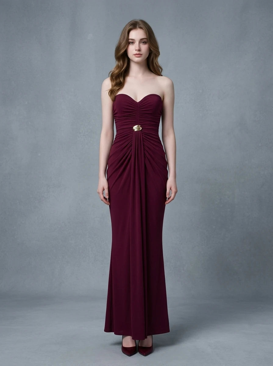 Woman in a burgundy strapless ruched maxi dress with gold waist embellishment, standing against a grey backdrop.