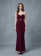 Woman in a burgundy strapless ruched maxi dress with gold waist embellishment, standing against a grey backdrop.