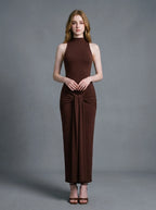 Woman wearing a long brown sleeveless mock neck dress with a knotted waist detail.