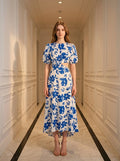 Woman wearing a white and blue floral print midi dress with puff sleeves and a cut-out waist, standing in a hallway.