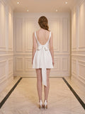 Back view of a woman in a white satin fit and flare dress with a square neckline and a large bow detail at the back.