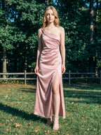 Woman in a dusty rose satin one-shoulder maxi dress with a high slit, standing in a grassy outdoor area with trees in the background.