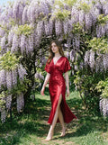 Woman in a red satin V-neck dress with kimono sleeves and a high slit, walking through a wisteria archway