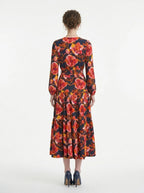 Back view of a woman in a navy blue long-sleeved midi dress with a red and yellow floral print, showcasing the tiered skirt and wrap silhouette.