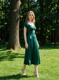 Woman standing in a park wearing a green satin midi dress with puff sleeves and a square neckline.