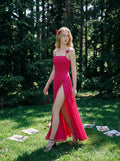 Woman in a bright pink ruched maxi dress with spaghetti straps and butterfly details, walking in a sunlit forest clearing.