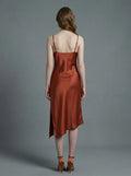 Back view of a woman in a rust-colored satin slip dress with spaghetti straps and an asymmetrical hemline, standing against a grey background.