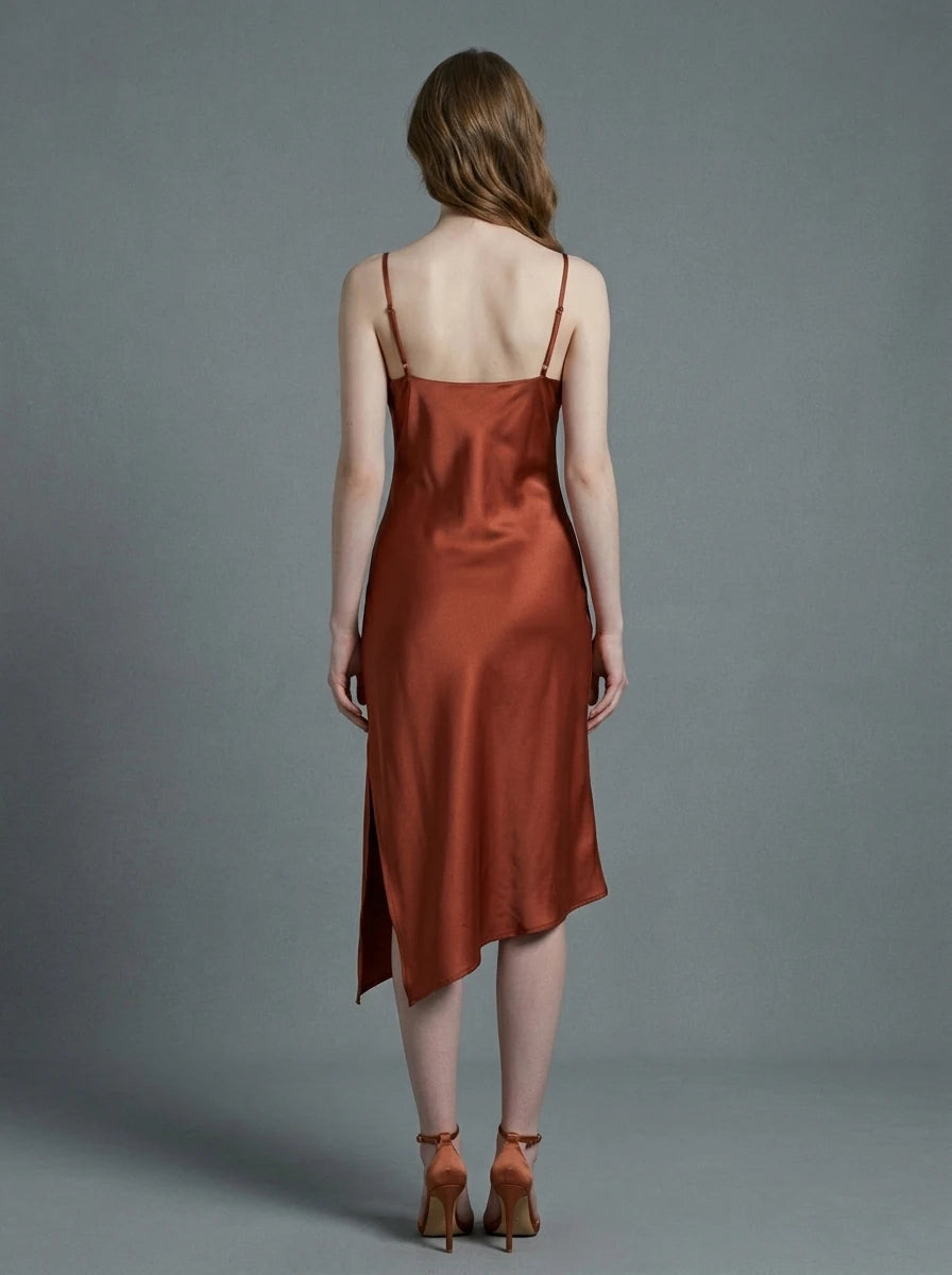 Back view of a woman in a rust-colored satin slip dress with spaghetti straps and an asymmetrical hemline, standing against a grey background.