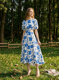 Woman wearing a white and blue floral print midi dress with puff sleeves and a cut-out waist, standing in a park.
