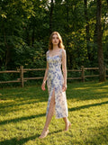 Woman wearing a floral print midi dress with tie shoulder straps and a thigh-high slit, walking in a grassy park.