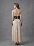 Rear view of a woman in a cream satin maxi dress with black lace detailing at the waist and a halter neckline, showing the open back and tie closure.