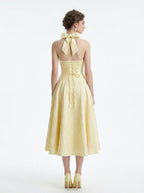 Back view of a pale yellow jacquard halter neck midi dress with a bow detail at the neck and a corset back.