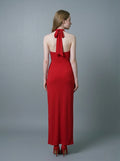 Back view of a woman in a red, floor-length, halter neck evening gown with a twisted waist detail and a high side slit, standing against a gray backdrop.