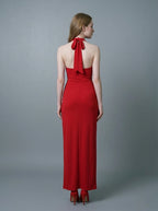 Back view of a woman in a red, floor-length, halter neck evening gown with a twisted waist detail and a high side slit, standing against a gray backdrop.