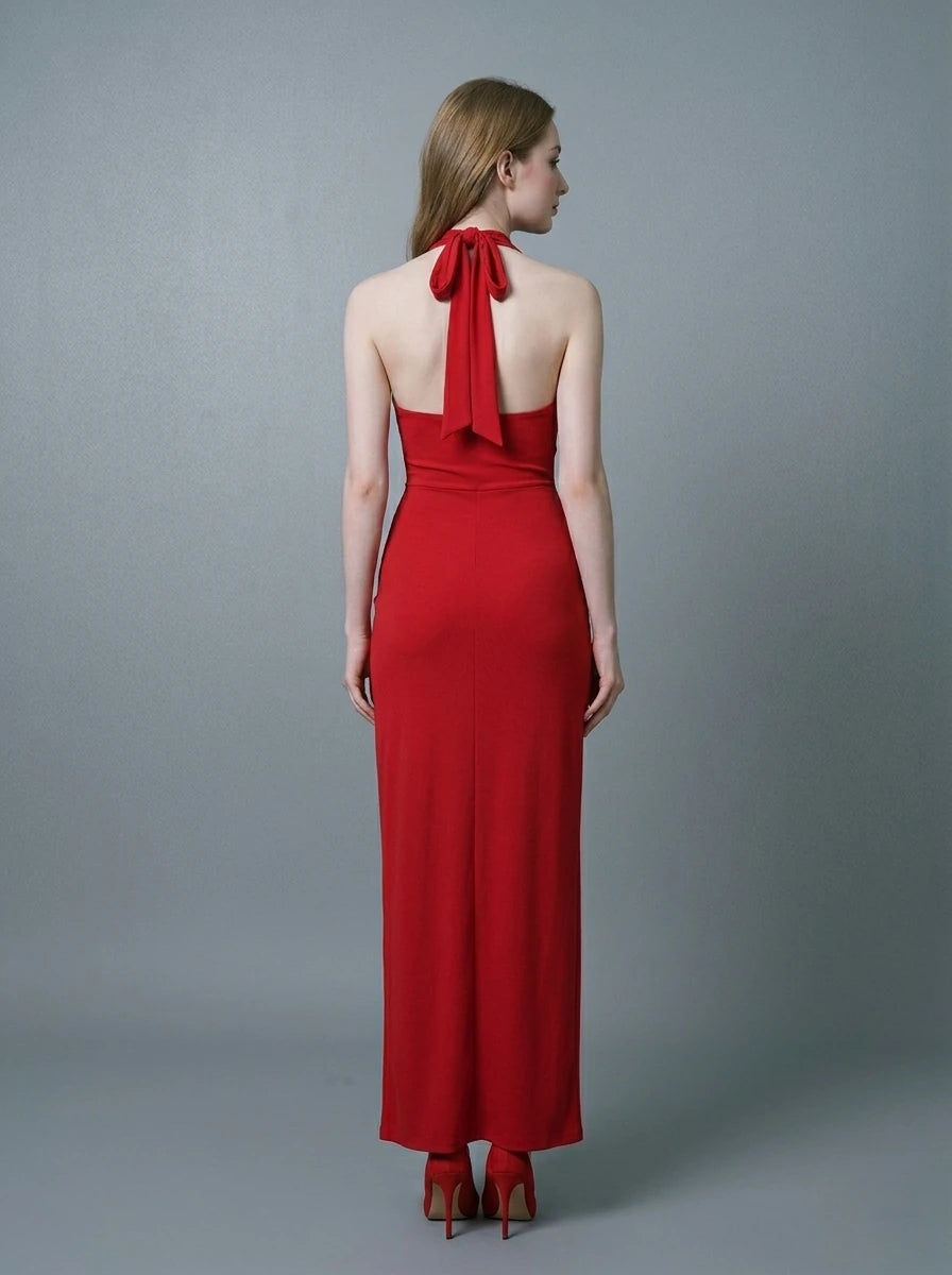 Back view of a woman in a red, floor-length, halter neck evening gown with a twisted waist detail and a high side slit, standing against a gray backdrop.