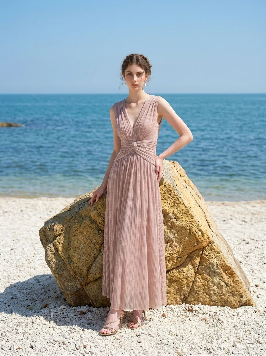 woman in a rose gold metallic a-line maxi dress with a v-neckline and ruched empire waist standing by a rock at the beach