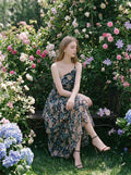 Woman sitting on a bench in a garden wearing a floral print one-shoulder chiffon dress with a tiered, asymmetrical ruffle skirt.