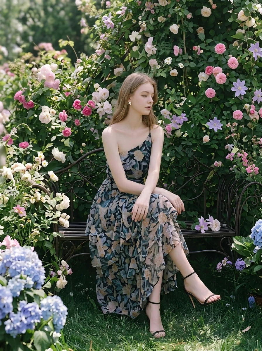 Woman sitting on a bench in a garden wearing a floral print one-shoulder chiffon dress with a tiered, asymmetrical ruffle skirt.