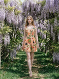 Woman in a peachy-pink, fruit and coffee cup print dress with a scoop neck and sleeveless design, walking through a garden path.