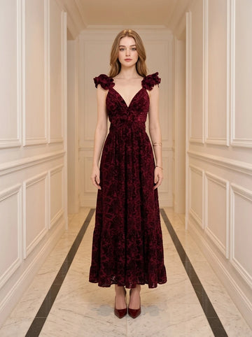 Woman wearing a burgundy velvet burnout A-line maxi dress with a V-neckline and ruffled cap sleeves, standing in a hallway.