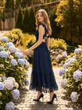 Woman walking on a cobblestone path surrounded by flowers, wearing a navy blue polka dot midi dress with an open back and criss-cross straps.