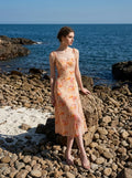 Woman in a peach floral print midi dress with side slit leaning against a rock on a rocky shore
