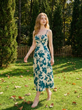 Woman in a cream and teal floral print midi dress with spaghetti straps and a tie-front bodice, walking in a grassy area with trees in the background.