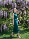 Woman in a dark green sleeveless halter neck sheath dress with ruched detailing and a high slit, walking outdoors under a wisteria tree