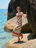 Woman standing on a rocky outcrop by the ocean, wearing a white maxi dress with a red floral print and red trim.