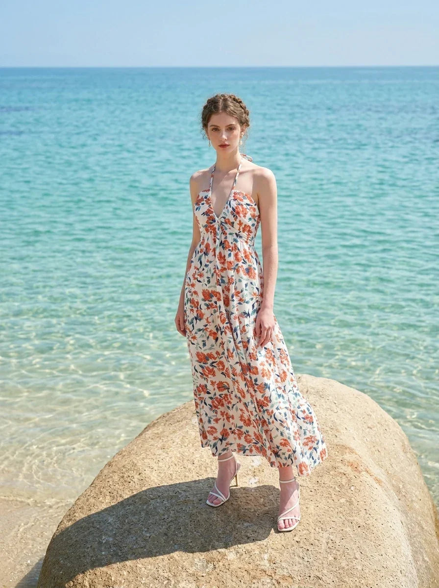 Woman standing on a rock in the ocean wearing a white floral print maxi dress with a halter neckline.
