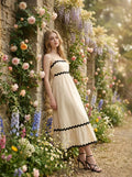 Woman leaning against a stone wall covered in flowers, wearing a cream-colored midi dress with black trim.