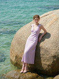 Woman in a lilac midi dress with a V-neckline and sleeveless design, leaning against a large rock by the ocean.