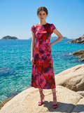 Woman with hand on hip on a rocky coastline, wearing a pink and red floral print satin ruched midi dress.