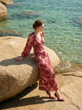 Woman sitting on a rock by the sea wearing a long, floral print, V-neck dress with long balloon sleeves.