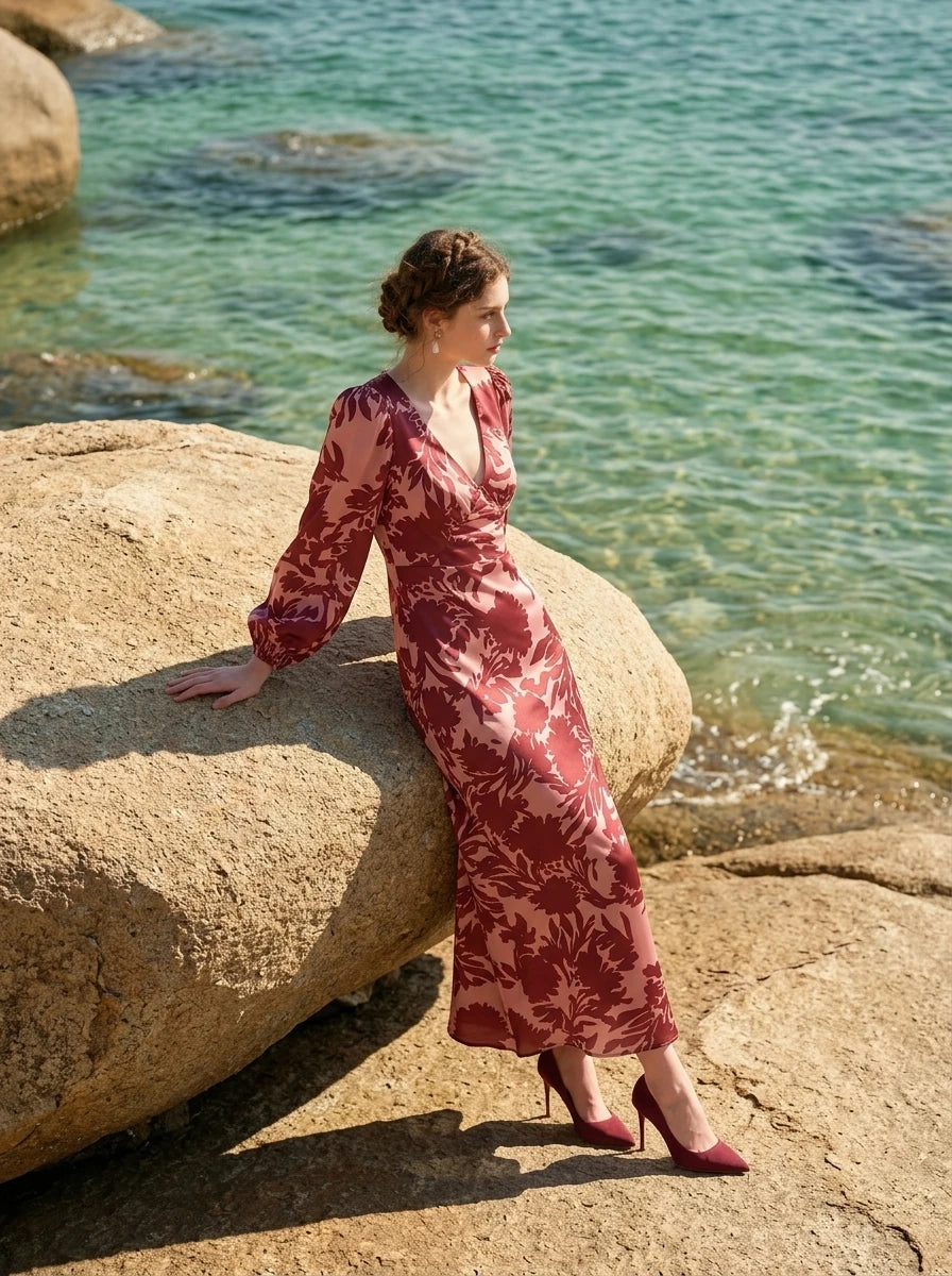 Woman sitting on a rock by the sea wearing a long, floral print, V-neck dress with long balloon sleeves.