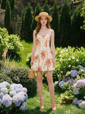 Woman wearing a white floral print A-line dress with a sweetheart neckline and adjustable shoulder straps, holding a straw hat and a basket bag, standing in a garden.