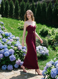 Woman wearing a floor-length, strapless burgundy gown with ruching and gold embellishment, walking on a stone path in a garden with hydrangeas.