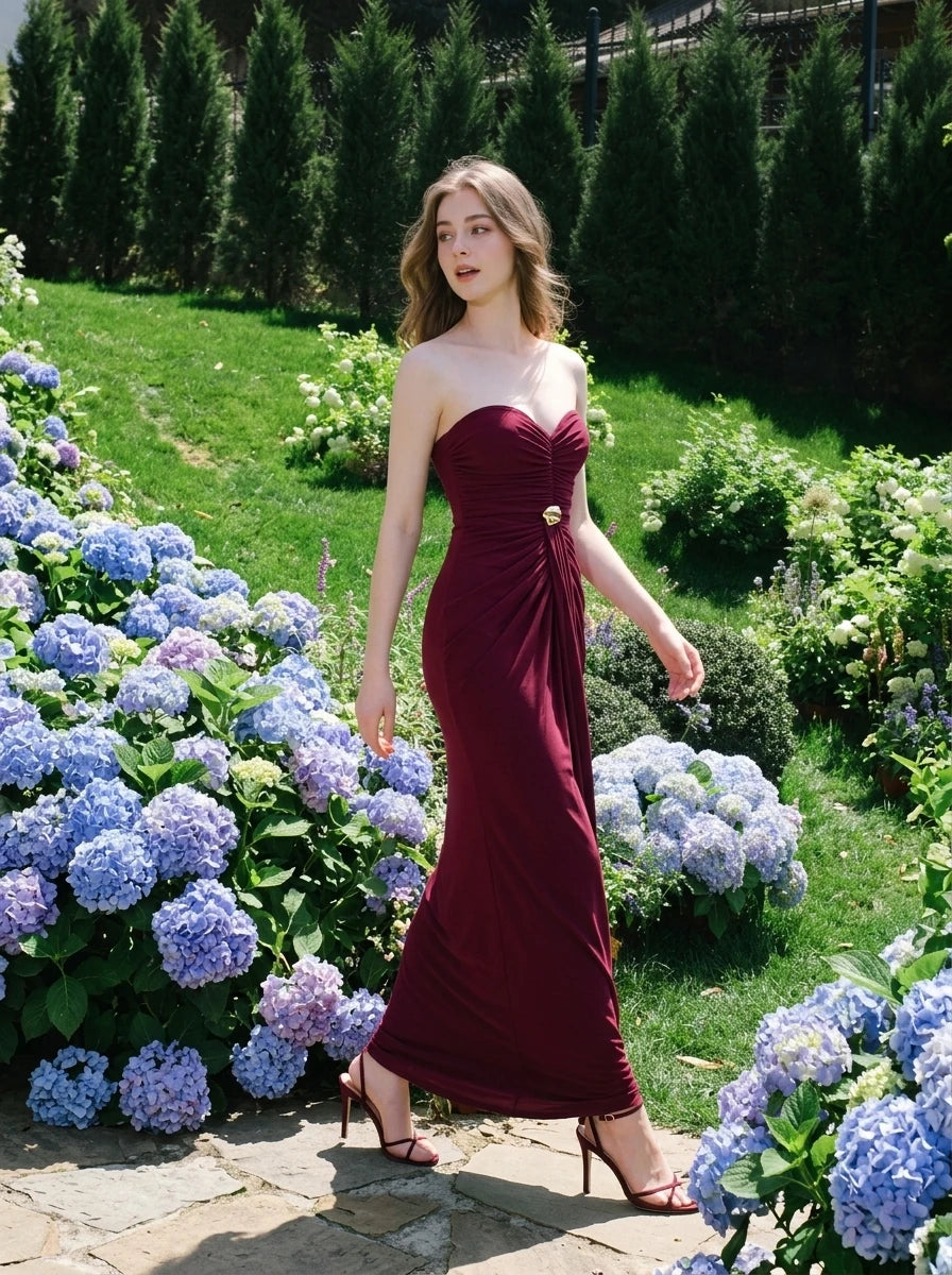 Woman wearing a floor-length, strapless burgundy gown with ruching and gold embellishment, walking on a stone path in a garden with hydrangeas.