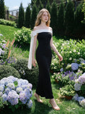 Woman in a black off-the-shoulder mermaid dress with a white draped accent on the shoulder, walking through a garden with hydrangeas and other flowers.